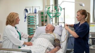 Älterer Herr im Krankenbett mit einer Ärztin und einer Krankenschwester neben dem Bett