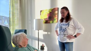 Eine ältere Frau sitzt mit verschränkten Armen auf einem Sessel und eine jüngere Frau steht ihr gegenüber